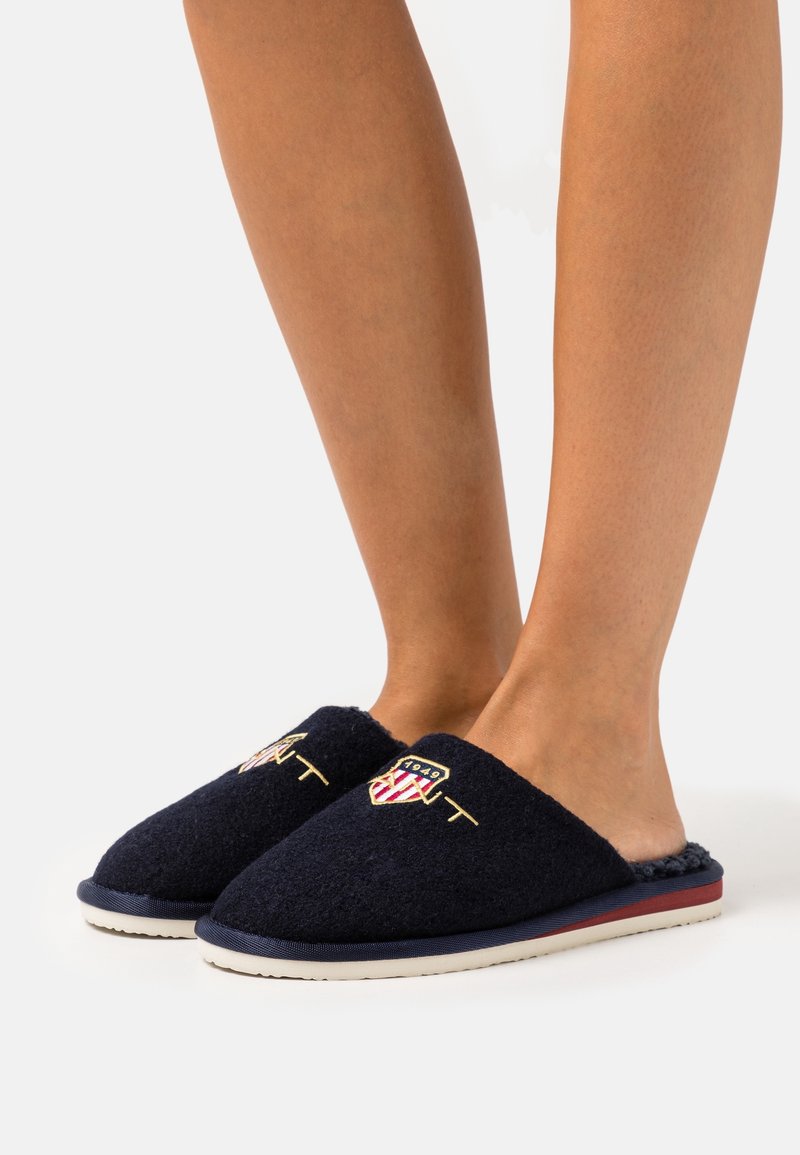 Pieds portant des chaussons bleu marine sans lacets avec un blason brodé en rouge, blanc et or ainsi que des lettrages, debout sur une surface blanche.