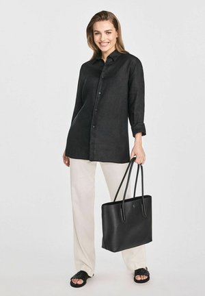 Kobieta uśmiechająca się, nosząca luźną czarną koszulę na guziki, białe spodnie, czarne plecione sandały i trzymająca dużą czarną torbę.