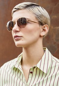 Runda solglasögon med bruna linser och guldmetallramar. Personen bär en randig grön- och vit skjorta med burgundy-accenter.