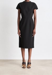 Femme portant une robe noire ajustée jusqu'aux genoux avec des manches courtes, et des sandales à talons ouvertes multicolores, debout devant un fond blanc.