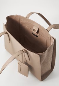 Beige tote bag gemaakt van gestructureerd leer met een gladde suède binnenkant, voorzien van twee handgrepen en een klein afneembaar label.
