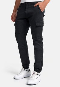 Bărbat purtând pantaloni cargo negri cu buzunare laterale și adidași albi Reebok, stând cu o mână în buzunar.