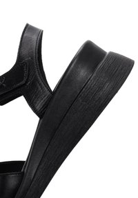 Sandalo nero con tomaia in pelle texturizzata, ampia cinghia e suola spessa, leggermente curvata. Presenta una chiusura a velcro per una facile calzata.