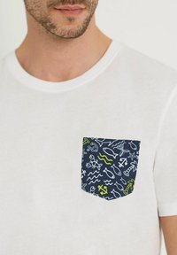 Valkoinen puuvillapaitoja, jossa on tummansinisessä taskussa värikkäitä meriteemaisia kuvioita, kuten kaloja, aaltoja ja ankkureita valkoisena ja limenvihreänä.