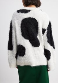 Osoba w puszystym białym swetrze z dużymi czarnymi abstrakcyjnymi plamami i ciemnozielonymi spodniami, pokazana od tyłu.