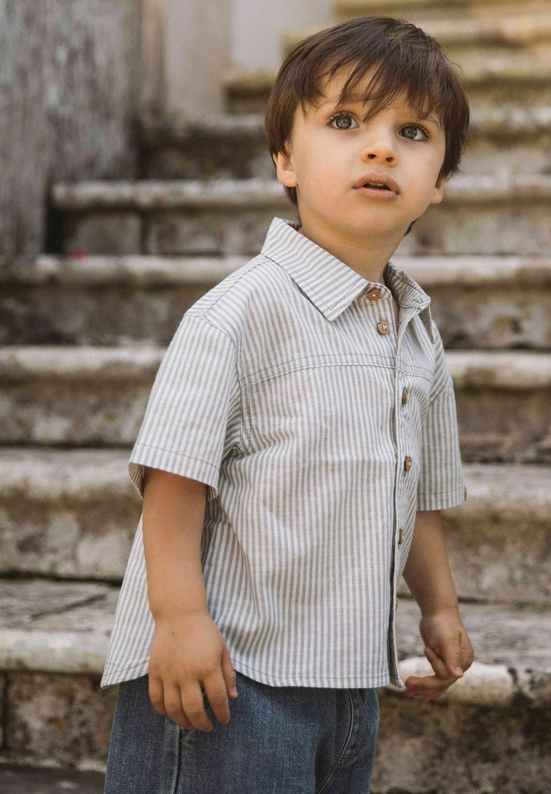 Jeune garçon aux cheveux bruns courts portant un t-shirt rayé à manches courtes et un jean, debout devant des marches en pierre en plein air.