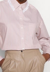 Camisa de rayas rosas y blancas de gran tamaño con un cuello blanco bordado, combinada con pantalones de cintura alta beige y bolsillos acentuados.