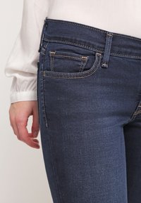 Vaqueros de mezclilla azul oscuro con un ajuste slim, que presentan un clásico diseño de cinco bolsillos, costuras visibles y una textura suave.