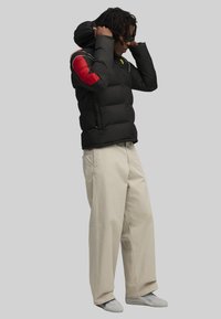 Zwarte puffer jacket met rode accenten op de schouder, met capuchon, in een boxy vorm. Gecombineerd met lichtbeige wijde broek en grijze sneakers.