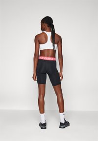 Fekete, testhezálló rövidnadrág, pink Nike logóval a derékpántján, fehér sportmelltartóval párosítva. A téma hátulról látható, álló helyzetben.
