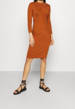 Femme portant une robe en maille ajustée, à manches longues, longueur genou, couleur rouille orange, et des sandales plates noires à lanières, debout devant un fond uni.