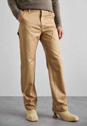 Persona vestida con pantalones beige de pierna recta con detalles de costura frontal, combinados con botas beige de ante con tacón y un suéter marrón.