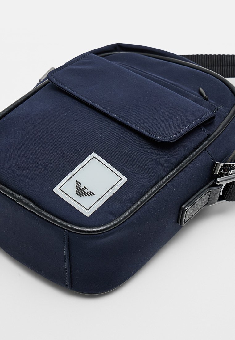 Bolso bandolera de nailon azul marino con un bolsillo frontal con solapa, ribete negro y un parche con el logo de la marca. Cuenta con una correa ajustable para llevar.