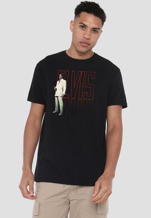 Junger Mann trägt schwarzes T-Shirt mit großem roten Schriftzug "ELVIS" und einem ganzflächigen Bild von Elvis Presley im weißen Anzug, der ein Mikrofon hält.