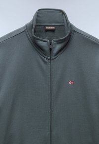 Lynlåsjakke i mørkegrønt stof med høj krave, central lynlås og lille norsk flagbroderi på venstre bryst.