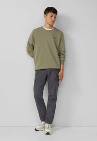 Giovane in felpa verde oliva, pantaloni cargo grigi e sneakers bianche, in piedi contro un muro grigio chiaro uniforme.