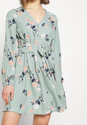 Kvinde iført en langærmet, lysegrøn kjole med pink og hvide blomsterprint og en bundet bue ved livet, stående foran en ensfarvet baggrund.