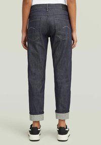 Jeans de mezclilla oscura con un ajuste entallado, puños enrollados, dos bolsillos traseros con costuras blancas y una etiqueta de logotipo negra en la cinturilla.