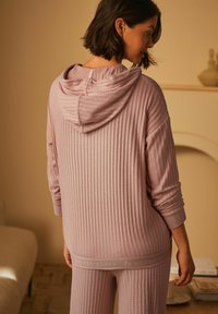 Rosa gerippter Kapuzenpullover mit einem strukturierten Muster, ausgestattet mit einer verstellbaren Kapuze und aufgerollten Ärmeln, kombiniert mit passenden Jogginghosen, die ein Logosband zeigen.