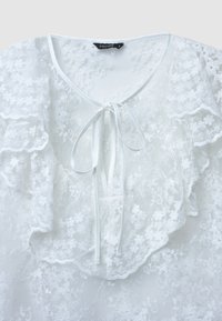 Blusa bianca in pizzo con bordi arricciati, un fiocco al collo e motivi floreali. La texture morbida e trasparente aggiunge un dettaglio delicato.
