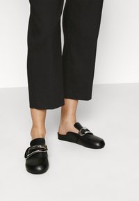 Zwarte leren muiltjes met een ronde neus en een open achterkant, met een grote metallic gesp aan de voorkant. Gepaarde met zwarte cropped broeken.