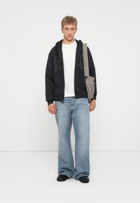 Svart hoodie med logotyp, vit t-shirt, ljusblå vida jeans, svarta skor och grå axelväska. Avslappnad, löst sittande stil.