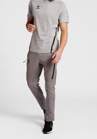Grå atletisk t-shirt og matchende tætsiddende joggers med sorte detaljer. Har et logo på brystet og sidelommer. Struktureret stof og snøre.