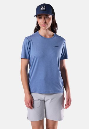 Kobieta ubrana w niebieską koszulkę Rossignol, jasnoszare szorty oraz ciemnoniebieską czapkę Rossignol, stojąca na tle jednolitego białego tła.