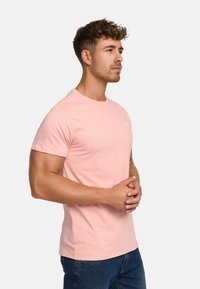 Short-sleeve, round-neck, light pink cotton t-shirt featuring a small white logo on the left chest, paired with dark blue jeans.