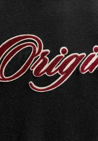 Gros plan sur une broderie rouge et blanche en script indiquant "Original" sur un fond de tissu noir.