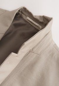Close-up of beige jacket collar showing stitching details and fabric texture on smooth, light background.