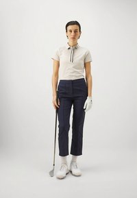 Πόλο πουκάμισο σε ανοιχτό μπεζ με μαύρο ριγέ γιακά, ναυτικές παντελόνες ραμμένες, λευκά παπούτσια γκολφ και λευκό γάντι που κρατάει μια ρόπαλο γκολφ.