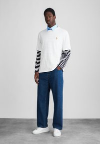 Mlad moški nosi slojen belo majico čez dolge črno-bele rokave, modro srajco, ohlapne modre kavbojke in bele superge.