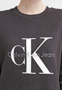Felpa nera in cotone, con un grande logo bianco "CK" e il testo "Calvin Klein Jeans" sottostante in un carattere pulito e moderno.