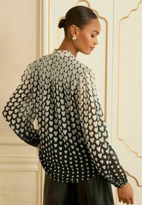 Blusa nera trasparente con motivi a cuore bianchi, maniche lunghe e retro arricciato; texture liscia e orlo arrotondato.