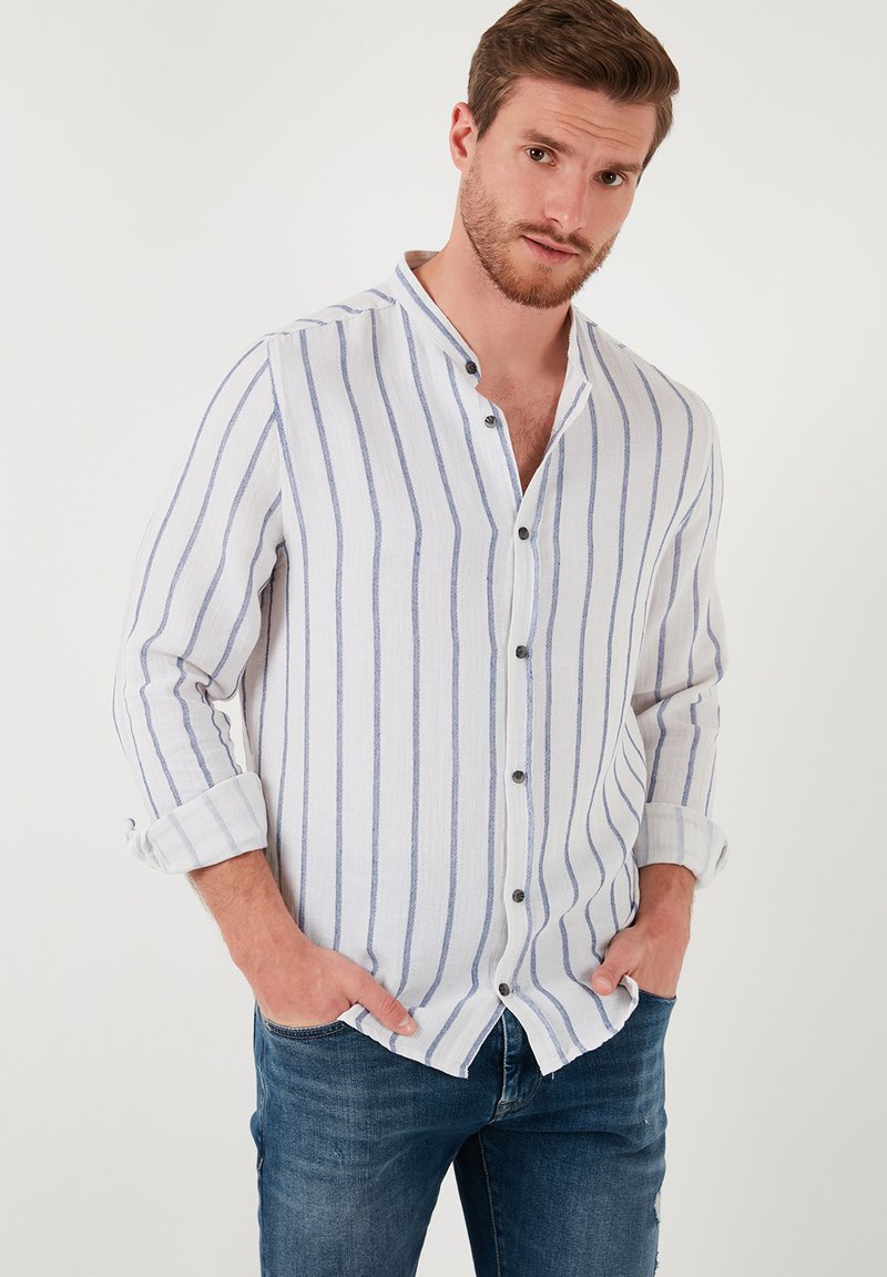 Camisa blanca de manga larga con rayas verticales azules, cuello mandarín y botones negros, combinada con jeans azules. Textura y diseño casual.