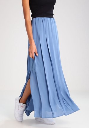 Femme portant une jupe maxi fluide bleu clair avec une fente latérale, associée à des baskets blanches et un top noir sans manches.