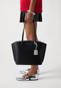 Borsa tote in pelle nera con hardware argentato e etichetta. Scolpita con un bordo superiore morbido, portata da una persona che indossa ballerine metalliche e una gonna plissettata.