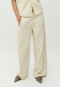 Pantalons larges beiges en tissu texturé, avec des poches avant et une fermeture à bouton. Portés avec des chaussures à bout pointu avec des accents de lanières.