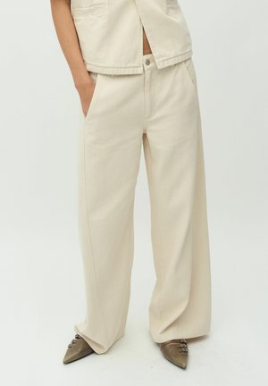 Beige weite Hosen aus strukturiertem Stoff, mit vorderen Taschen und Knopfverschluss. Getragen mit spitz zulaufenden Schuhen mit Riemenakzenten.