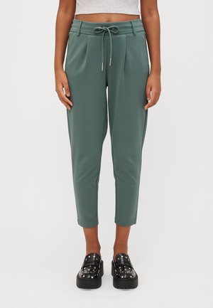 Pantalons verts effilés avec taille à cordon, dotés de plis et d'une texture lisse, associés à des mocassins à plateforme noirs avec des accents cloutés.