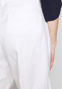 Vitt bomullstyg, smal passform, med en bakficka som har knappstängning. Slät textur, odekorerad design.