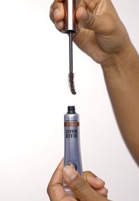 Schwarze Magic Mascara-Tube mit silberner Oberfläche und schwarzem Deckel. Bürste mit kurzen Borsten, die darüber gehalten wird. Hand, die den Tube greift, mit glatter Hautstruktur.