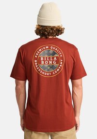 T-shirt couleur rouille avec un graphique circulaire comportant le texte "BILLA BONG" et des accents décoratifs. Fabriqué en coton doux.