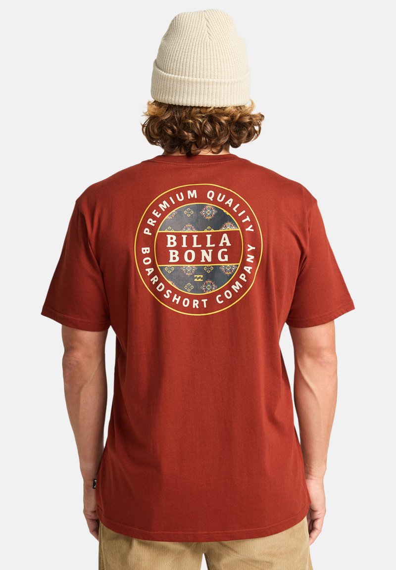 T-shirt couleur rouille avec un graphique circulaire comportant le texte "BILLA BONG" et des accents décoratifs. Fabriqué en coton doux.