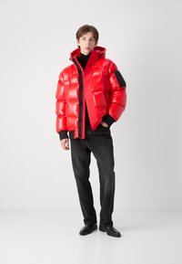 Rød puffjakke med en blank overflade, hætte, lynlås og sorte accenter. Bæres over en sort turtleneck og parret med mørke jeans.