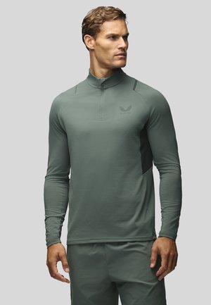 Homme portant un haut de sport vert moulant à manches longues avec une fermeture éclair au quart et un short assorti, debout devant un fond uni.