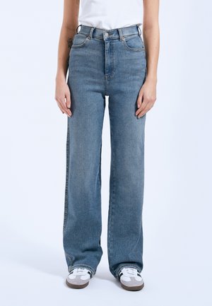 Persona che indossa jeans blu a vita alta con gamba dritta e sneakers bianche, in piedi davanti a uno sfondo chiaro e uniforme.