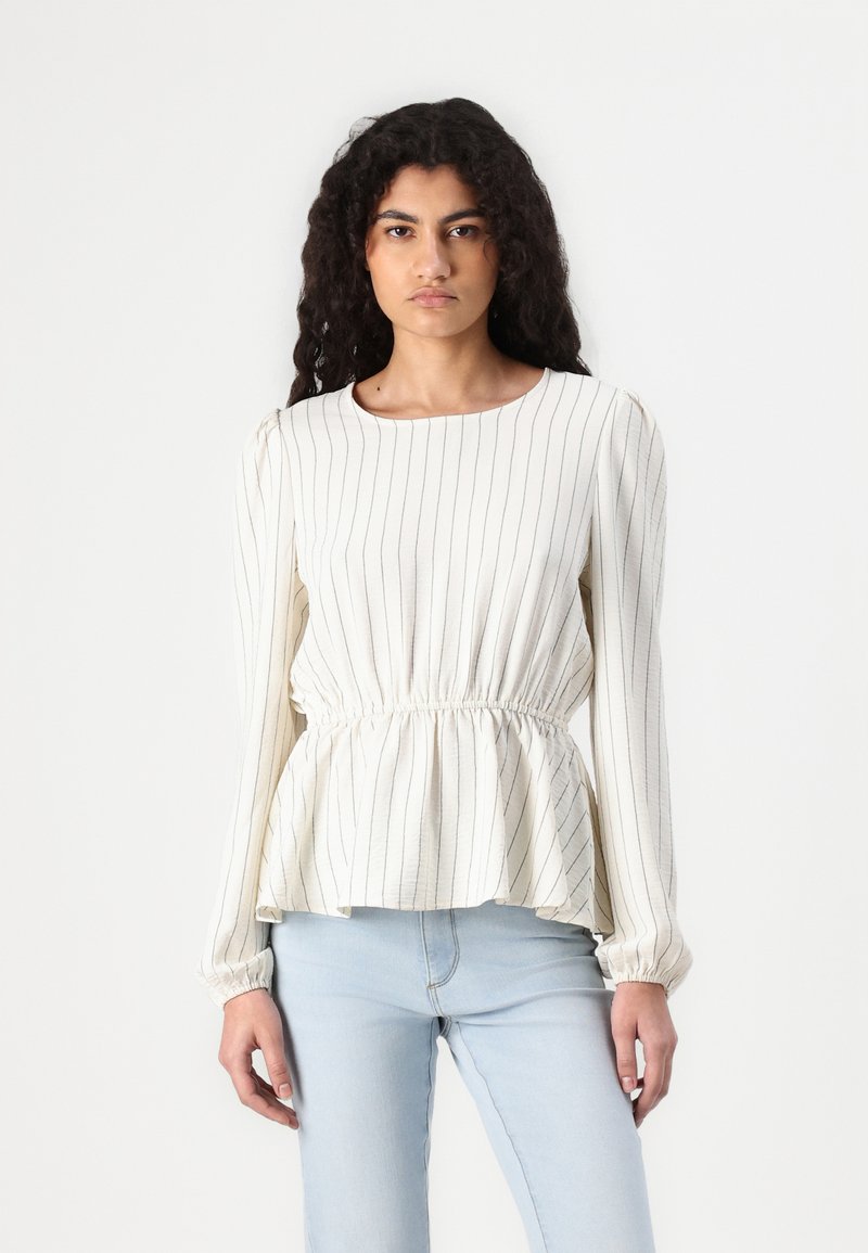Blouse peplum blanche à rayures, à manches longues, avec une taille élastique et un tissu doux. Associée à un jean bleu clair.
