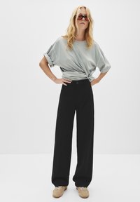 Vrouw met lang blond haar, die oranje getinte zonnebril draagt, een opgerolde grijze t-shirt die om haar taille is geknoopt, zwarte wijde broek en beige veterschoenen.